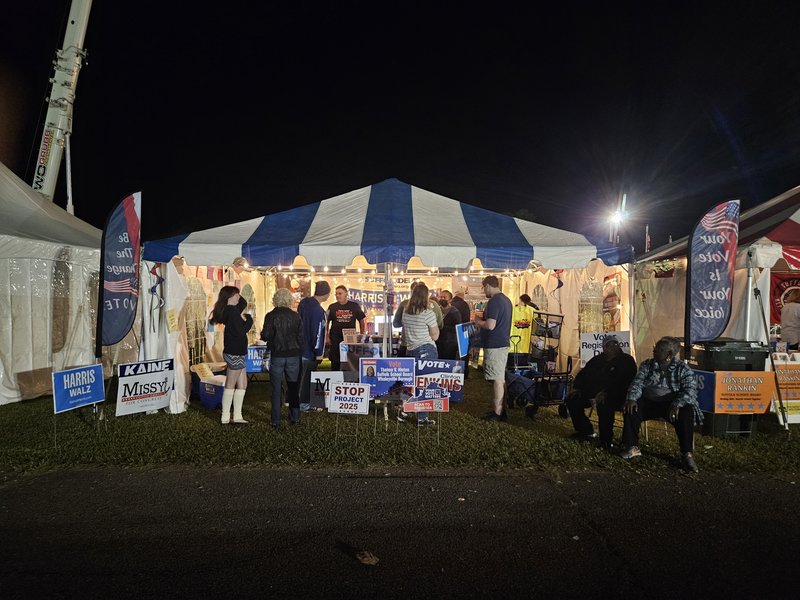About a dozen people in and around an event tent, lit from the inside, and decorated with United States of America symbols and colors, and with political campaign signs
