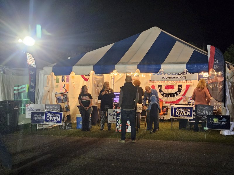 20 x 20 event tent, open in front.  Decorations include USA red-white-blue banners and bunting and candidate yard signs.  About a half-dozen people are in or in front of the tent. The sky is black and the tent is well lit